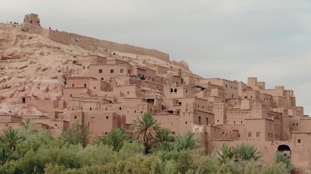 The ancient fortified city of Ait Benhaddou in Morocco stands tall against a clear sky. Tourists explore the historic ksar, a UNESCO World Heritage site.
