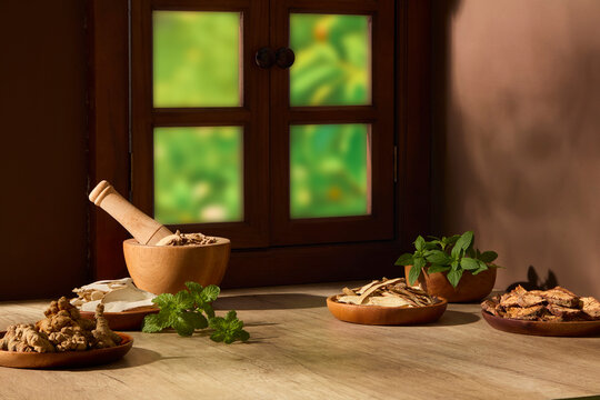 Rustic still life of dried herbs, roots, and natural ingredients arranged on wooden table near a window. Warm sunlight, traditional herbal atmosphere. Design product on blank surface