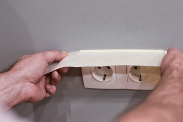 In a detailed close-up, a person uses strong sticky tape to seal off a wall power socket, ensuring it remains clean and paint-free while the surrounding area is being painted.