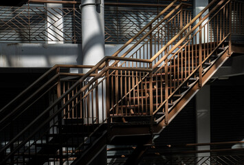 A prominent, industrial-style metal staircase climbs through a high-contrast environment, dramatically illuminated by natural light that carves sharp divisions of shadow and brilliance across structur
