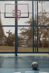 A worn basketball rests on the blue cement floor of an outdoor court with a basketball hoop, fiberglass backboard, and blue steel fence comprising the background. Copy space.