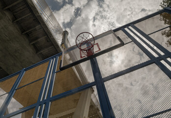 Through the steel grid fence, the eye is led past the orange metal basketball rim and net, up to the industrial ring road bridge and the dramatic sky beyond. Copy space.