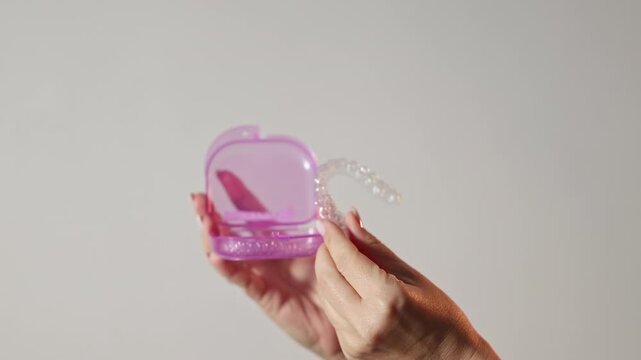 Woman uses dental trays for teeth whitening at home. Girl pours peroxide solution from syringe and applies on teeth, smiling to camera. Neutral background. Female holds trays in a purple case.