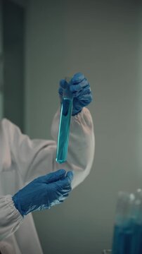 Close-up of a chemist's hands in blue gloves shaking a test tube with a transparent blue chemical liquid during a laboratory experiment