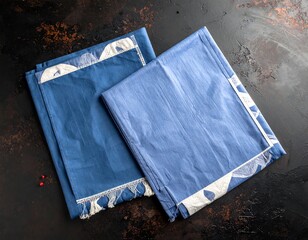 Two folded, blue fabric napkins with a white patterned border on a mottled brown and black surface