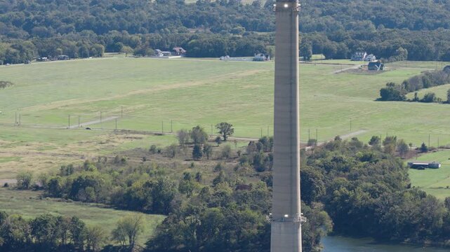 Tall smokestack rises above rolling farmland and patchwork fields under clear daylight conditions, tilt down to power plant operations