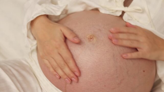 Pregnant woman lies on bed and applies cream on belly. Female in last month of pregnancy uses anti stretch mark cream from jar. She takes product with finger and gently massages abdomen. Body care rou