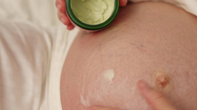 Pregnant woman lies on bed and applies cream on belly. Female in last month of pregnancy uses anti stretch mark cream from jar. She takes product with finger and gently massages abdomen. Body care rou