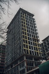 A tall building under construction, showcasing a modern architectural design with steel frames and scaffolding against a cloudy sky.