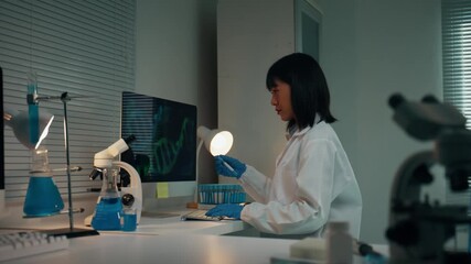 Young Asian woman in a lab coat and gloves meticulously examining a test tube at her desk. Professional working with dna and genetic research - Powered by Adobe