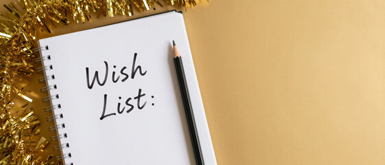 A white spiral notebook with Wish List written on it sits next to a black pencil and golden tinsel on a beige background. Christmas holiday planning.