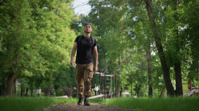 Man Walking Toward Camera Smiling With Backpack And Sunglasses, Confident Stride On Sunlit Path Flanked By Lush Trees, Close Perspective Captures Energy And Relaxed Outdoor Vibe, Cargo Pants