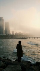 Man in trench coat and hat stares out at moody ocean near foggy city skyline