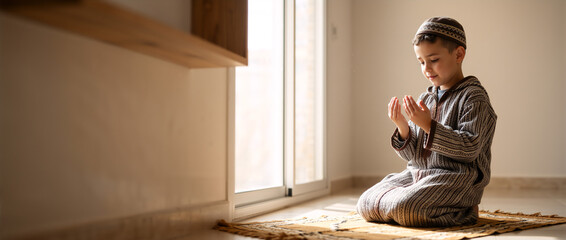 Young Muslim boy praying on a prayer rug at home. Child in traditional clothing performing Salah during Ramadan. Spiritual Islamic banner with copy space