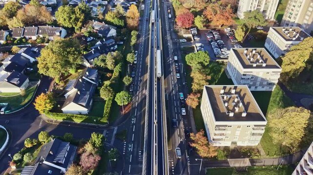 Top-down aerial drone view of a metro train and road traffic moving in parallel through a residential district of Rennes. Autumn colors line the urban infrastructure, France
