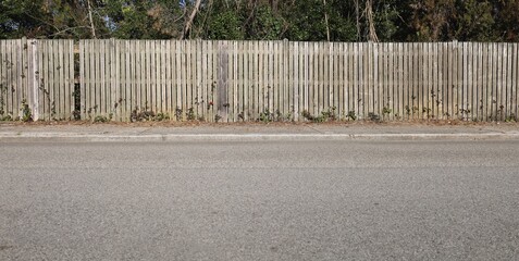 Weathered wooden fence at roadside. Trees on behind, sidewalk and street in front. Background for copy space.