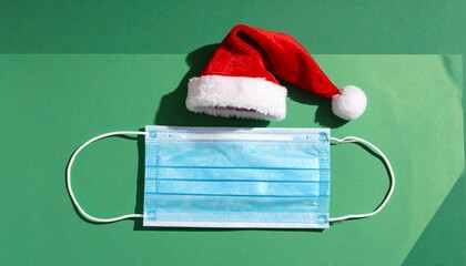 Blue medical face mask wearing red Santa hat on green background, creative Christmas health concept, minimalist flat lay, soft studio lighting, clean composition, festive message