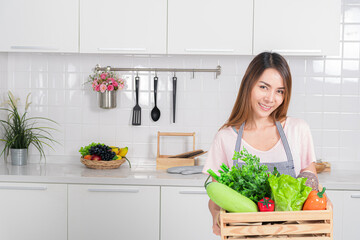 Smiling beautiful asian woman in an apron hold a wooden crate filled with organic fresh colorful vegetable promoting healthy eating and cooking.