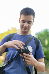Fototapeta premium A man is holding a camera and smiling. He is wearing a blue shirt and a backpack. The camera is attached to a strap around his neck