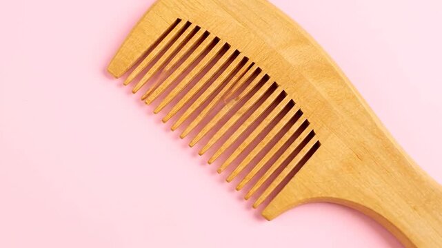 A wooden comb with closely spaced teeth on a solid light pink background. Comb positioned at a diagonal