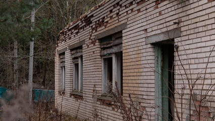 A brick building stands in a wooded area with broken windows and visible signs of decay. Weeds and plants grow around the base, showing signs of neglect over time.
