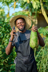 An African male farmer wearing a straw hat and black apron smiles while holding a green papaya and a mobile phone outdoors, standing in a lush farm environment surrounded by vegetation.