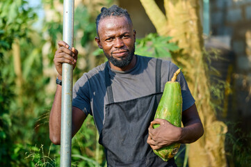 A Nigerian male farmer wearing an apron holds a ripe green papaya while standing outdoors beside a metal pole in a farm setting with trees and natural sunlight.