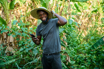 An African male farmer wearing a straw hat smiles while holding a mobile phone and resting a farming tool on his shoulder inside a lush banana plantation.