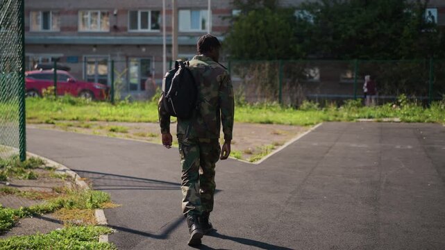 Military Figure Moves With Reflective Pace Toward Urban Crossroads And Fence, Soldier Equipped With Backpack Advances Slowly Towards City Junction Under Sunlight Near Fences And Apartment Buildings