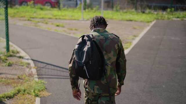 Black Soldier Walking Down Forked Path With Backpack And Camouflage, Back To Camera. Urban Crossroads And Sunlit Pavement Frame Decisive Movement Toward Future And New Direction. Wide Composition