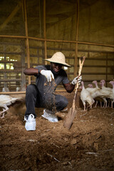 Young Nigerian male poultry farmer crouching inside an indoor turkey farm, wearing a straw hat and gloves while sprinkling soil beside a shovel as white turkeys stand nearby.