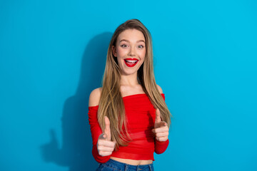 Smiling young woman with long hair pointing at viewer in red top against blue background, showcasing casual style and joy