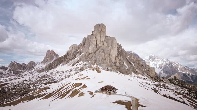 Aerial drone moves rightways past Ra Gusela peak and rifugio at snowy Passo Giau mountain pass in the Dolomites, Italy, under dramatic cloudy sky.