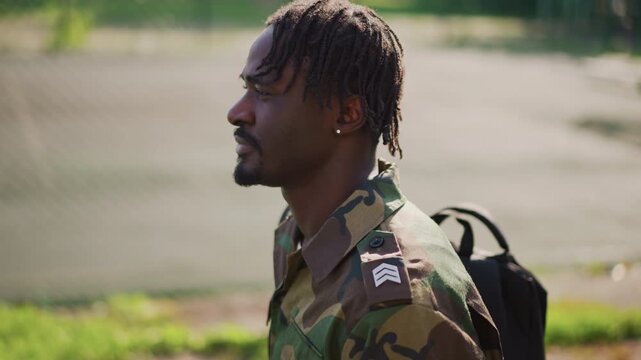 Reflective Soldier Walking, Veteran With Backpack Beside Fence, Quiet Moment Captured Of Military Veteran Reflecting, Contemplative Veteran In Camouflage Gear Walks Past Fence During Evening Hours