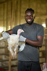 Young African male poultry farmer smiling while holding a white turkey inside an indoor farm, wearing gloves and a casual t shirt, standing in a livestock barn environment.