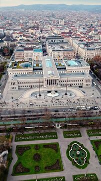 Aerial portrait drone shot panning left over the Austrian Parliament Building, revealing the Vienna cityscape with historic rooftops, streets, and light pedestrian and traffic under winter sunlight