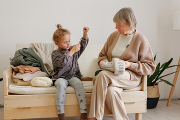 A grandmother watches as her granddaughter enthusiastically knits on a sofa. The room is bright and inviting, filled with yarn and cozy blankets