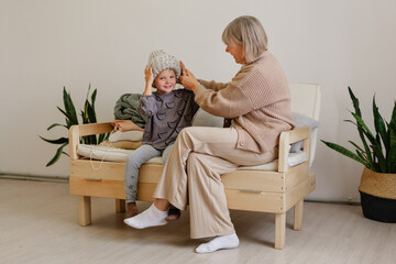 Obraz premium A grandmother smiles as she assists her young grandchild in wearing a knitted hat at home. They are surrounded by soft pillows and plants