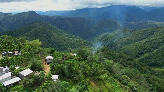 Onwards aerial surpassing rural mountain houses in Banaue, Ifugao, gliding toward rising smoke with vast, layered mountain ranges in the distance in Banaue, Ifugao, Philippines.