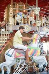 A young man is riding a carousel horse. The carousel is decorated with many lights and has a colorful design. The boy is wearing a yellow shirt and shorts © Masakazu Tokashiki