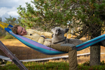 Woman relaxing in hammock with dog wearing outdoor clothing in forest nature