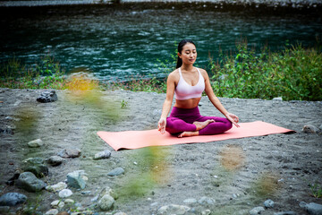 Young Japanese woman meditating in lotus pose during outdoor yoga by a river, calm sustainable lifestyle concept with mindfulness and mental wellbeing