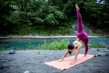 Asian woman practicing advanced yoga balance pose outdoors by a river, strength and mental wellness lifestyle in nature