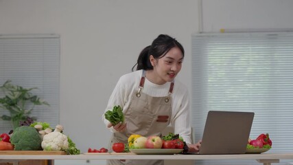 Pretty Asian woman wearing an apron live streaming a cooking class on her laptop. She is showing fresh lettuce to the camera
