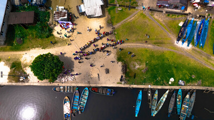 Aerial view of boats lining the dark water's edge contrasting with the vibrant green vegetation and the sandy shore bustling with figures in Soku Community, Abonnema, Port Harcourt, Nigeria.