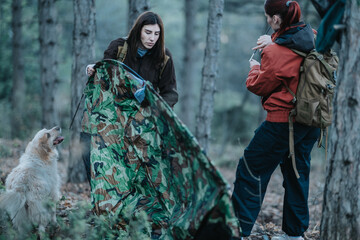 Two hikers and a dog in a forest, setting up a camo tent during a camping outing. © qunica.com