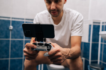 Close-up view of bearded young adult man focusing on mobile gaming using smartphone and gamepad while sitting on toilet, portraying modern habits and technology integration in daily life.