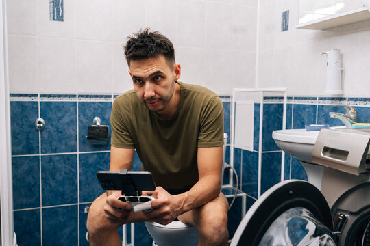 Man playing mobile video games with controller attached to smartphone, sitting on toilet in residential bathroom, showing digital addiction and modern habits. - Powered by Adobe