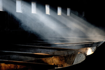 Solemn interior of ancient galley ship illuminated by mysterious shafts of light before naval battle, showing wooden oars in dark smoky hull, evoking mood of tension and anticipation before war