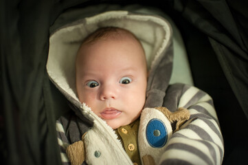 An infant is lying in a stroller looking up with a shocked expression. The baby wears a striped hoodie and a buttoned shirt. The background appears dark.